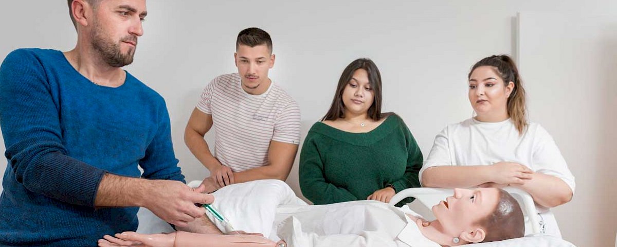 Ein Pflegeschüler verabreicht eine Spritze an einer Pflegepuppe. Drei weitere Schüler schauen dabei zu