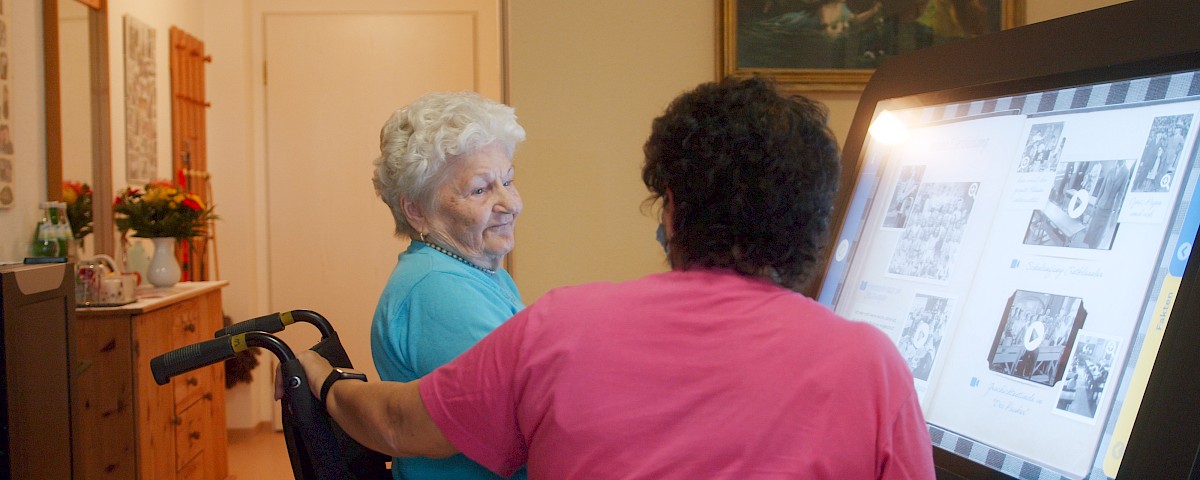 Eine ältere Dame sitzt vor einem Care Table, ein digitaler Aktivitätstisch für die Altenhilfe, 43-Zoll-Touchdisplay (ca. 109 cm), das in einen Tisch eingelassen ist.