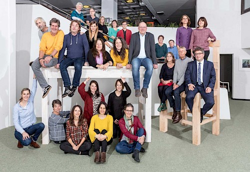 Lehrerkollegium Evang. Fachschule für Sozialpädagogik Lehrerinnen und Lehrer sitzen auf einem überdimensional großen Tisch und einem Stuhl